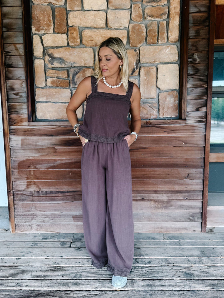 A woman modeling a mauve-colored sleeveless jumpsuit with wide-leg pants, standing on a rustic wooden porch in front of a stone wall.