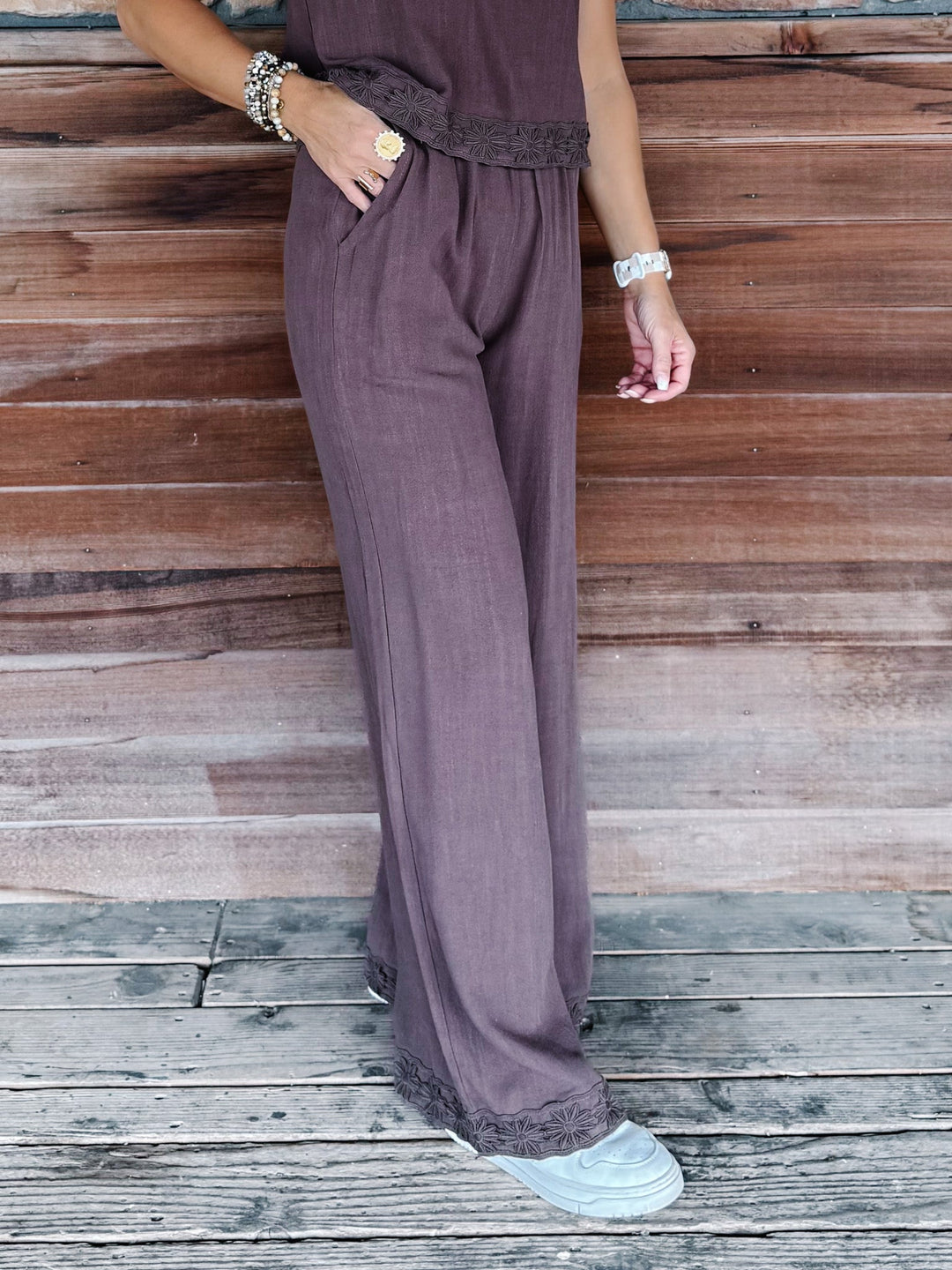 A person wearing wide-leg mauve-purple pants with a matching top and white sneakers, standing on a weathered wooden deck against a wooden wall.