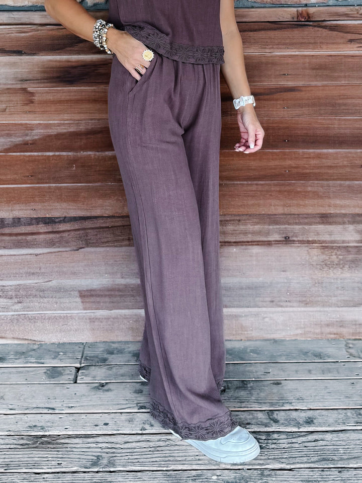 A person wearing wide-leg mauve-purple pants with a matching top and white sneakers, standing on a weathered wooden deck against a wooden wall.