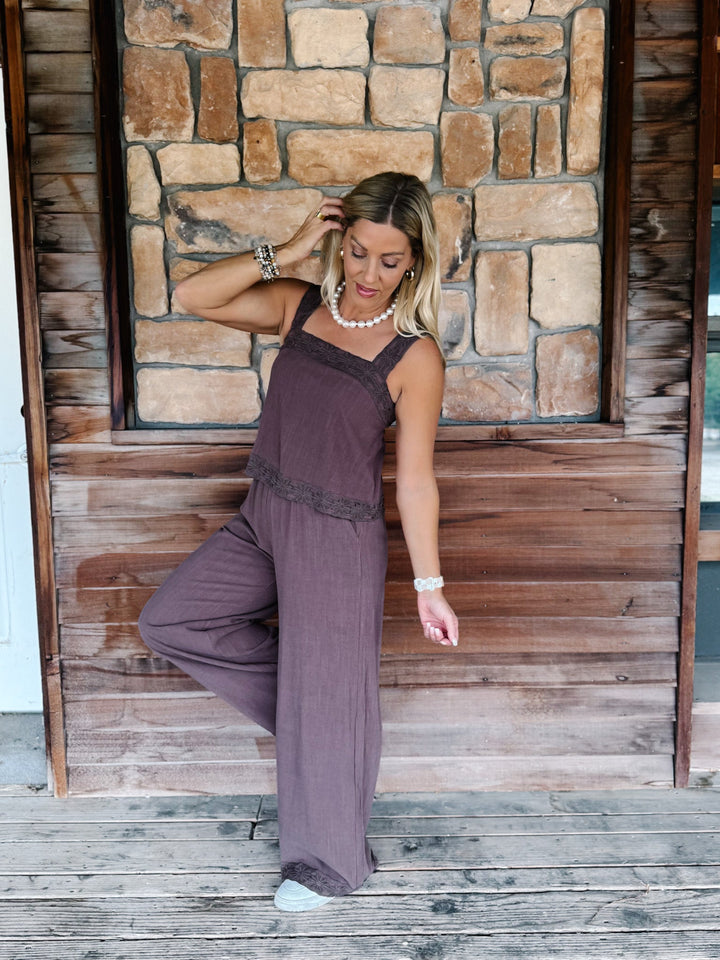 A woman modeling a mauve-purple sleeveless jumpsuit with lace trim, posing on a rustic wooden porch with a stone wall background.