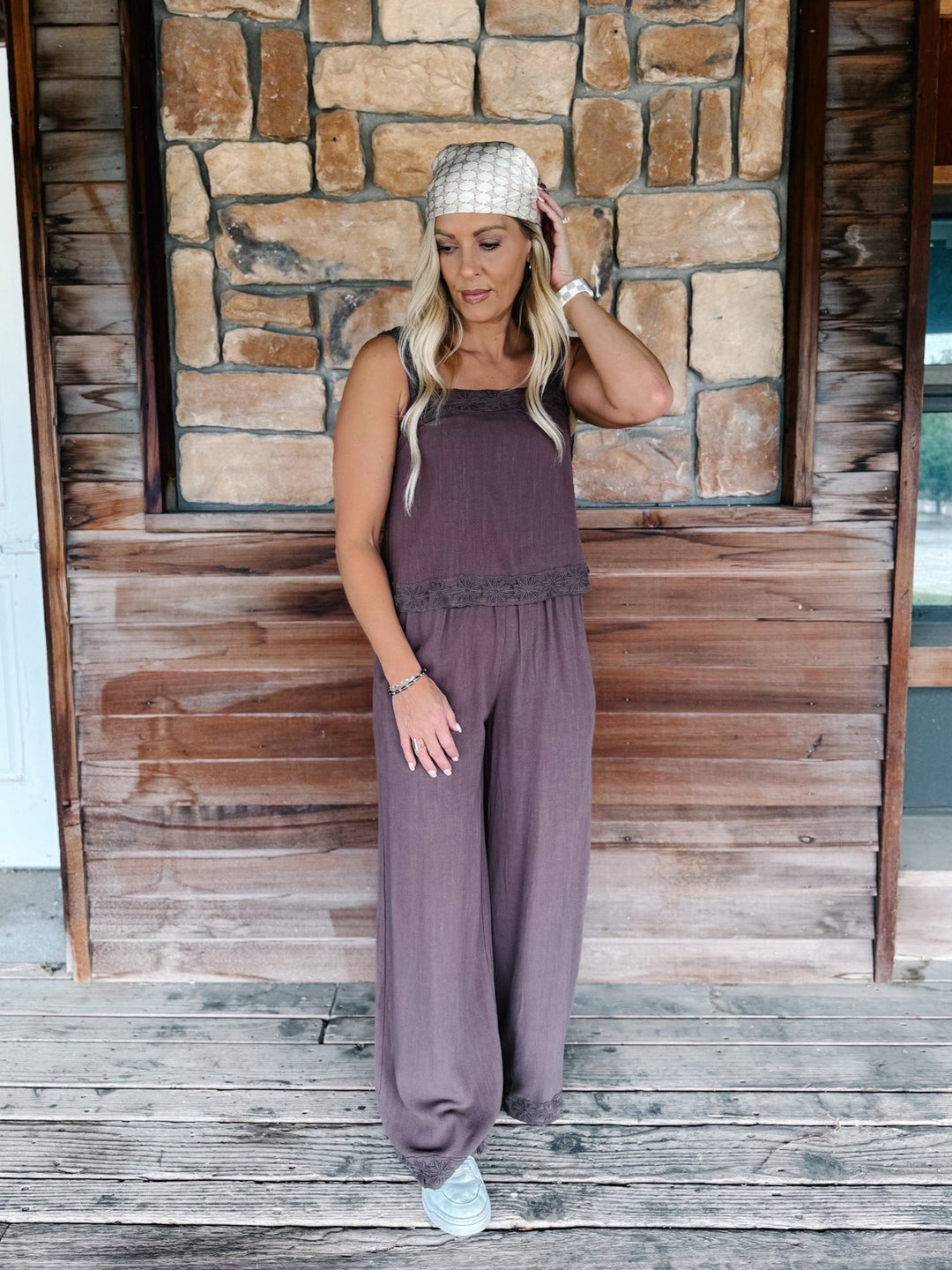 A woman stands on a rustic wooden porch in front of a stone wall, wearing a sleeveless mauve-purple jumpsuit with wide legs and a textured knit head covering.