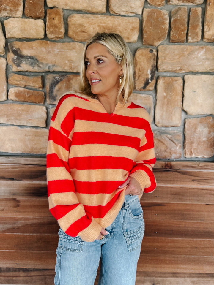 A woman wearing a red and beige striped knit sweater with a split neckline, paired with light blue jeans, standing in front of a rustic stone wall and wooden floor.