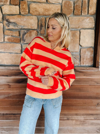 A woman wearing a red and beige striped knit sweater and light blue jeans, standing in front of a rustic stone wall and wooden bench.
