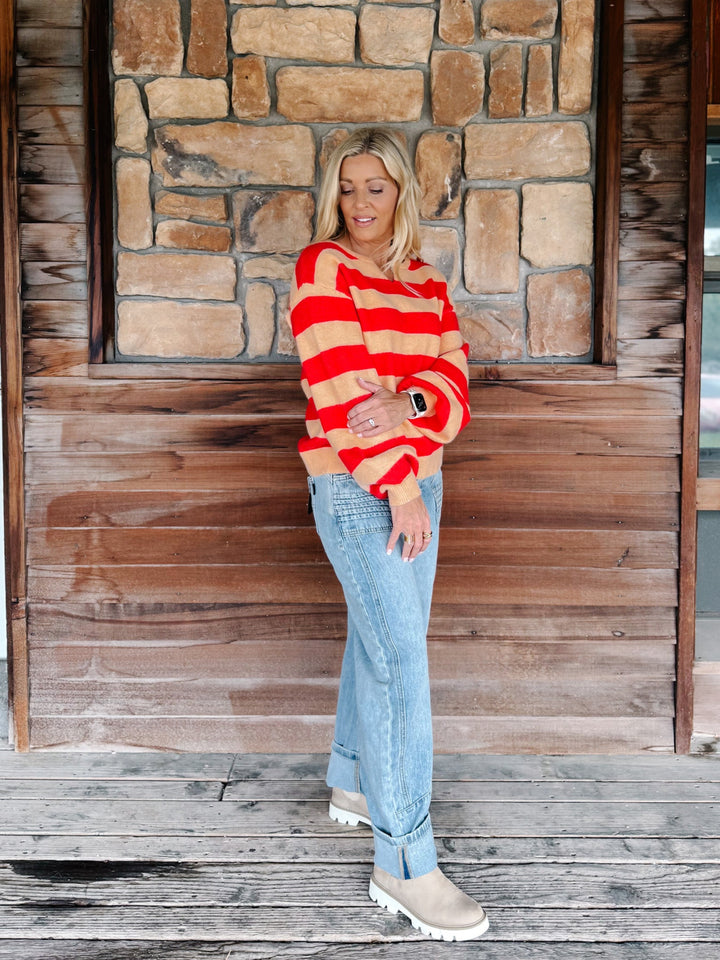 A woman stands on a rustic wooden porch in front of a stone wall, wearing a red and beige striped knit sweater, light blue jeans, and beige boots.