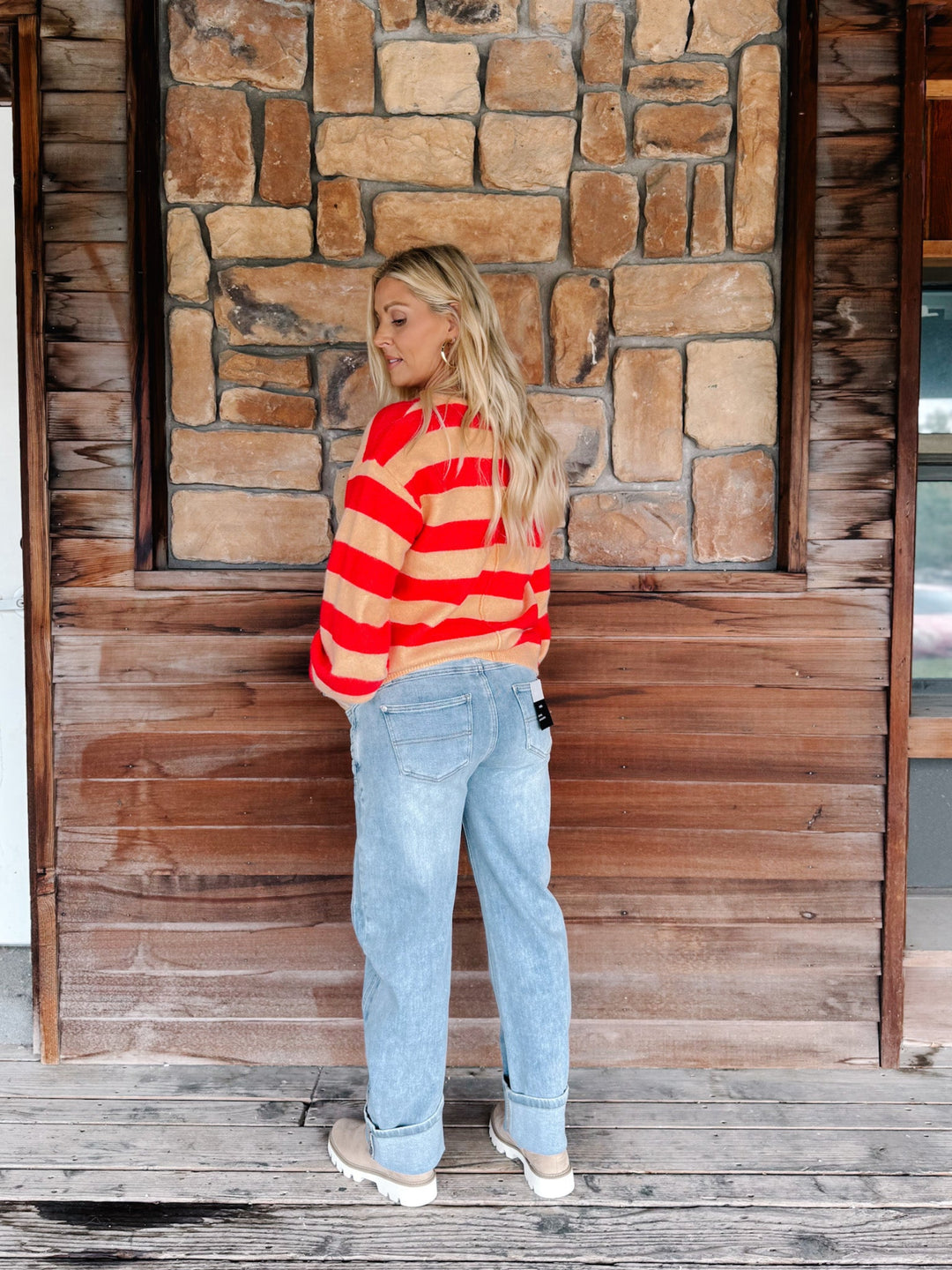 A woman stands on a wooden porch wearing a red and orange striped long-sleeve crop top, light blue jeans, and beige sneakers, set against a rustic stone and wood wall.