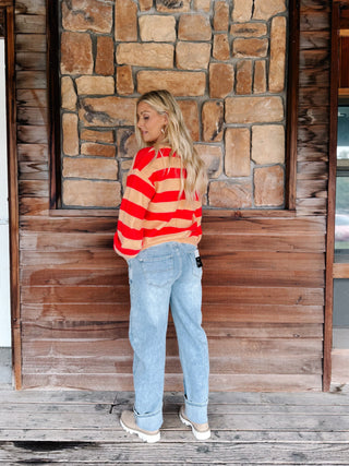 A woman stands on a wooden porch wearing a red and orange striped long-sleeve crop top, light blue jeans, and beige sneakers, set against a rustic stone and wood wall.