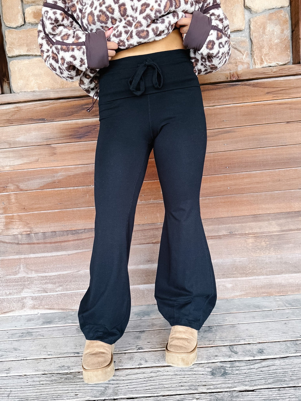 A person wearing black wide-leg harem pants with a drawstring waist, paired with a leopard-print sweatshirt and tan platform shoes, standing in front of a rustic wooden wall.