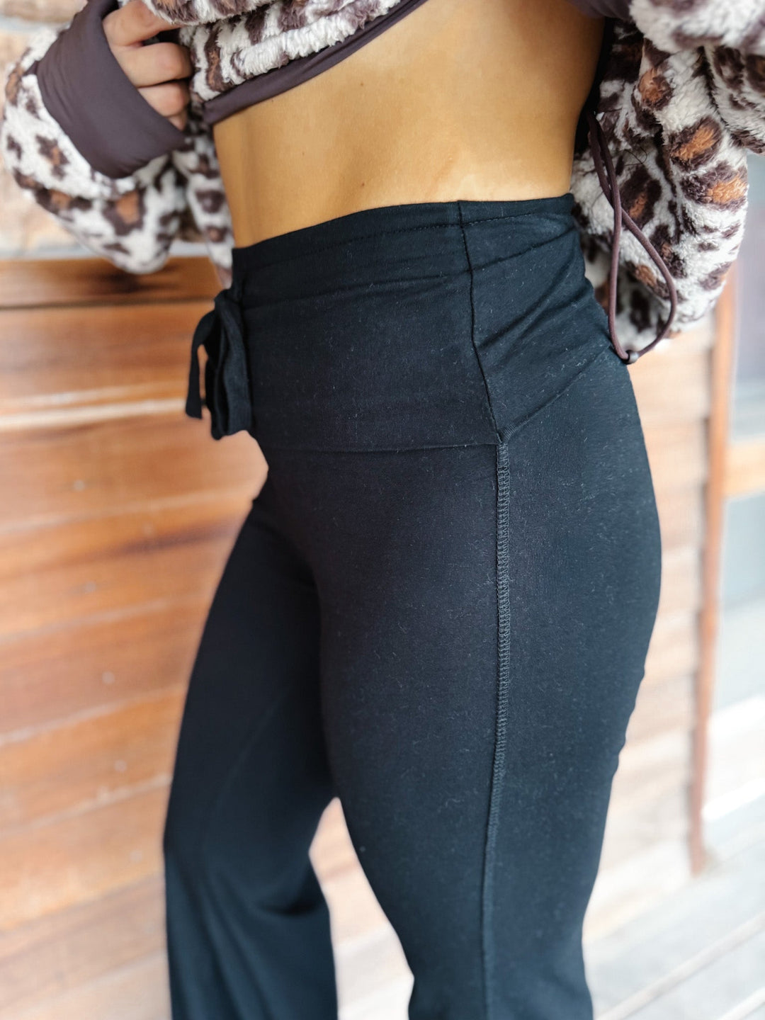 A person wearing black wide-leg harem pants with a drawstring waist, paired with a leopard-print sweatshirt and tan platform shoes, standing in front of a rustic wooden wall.