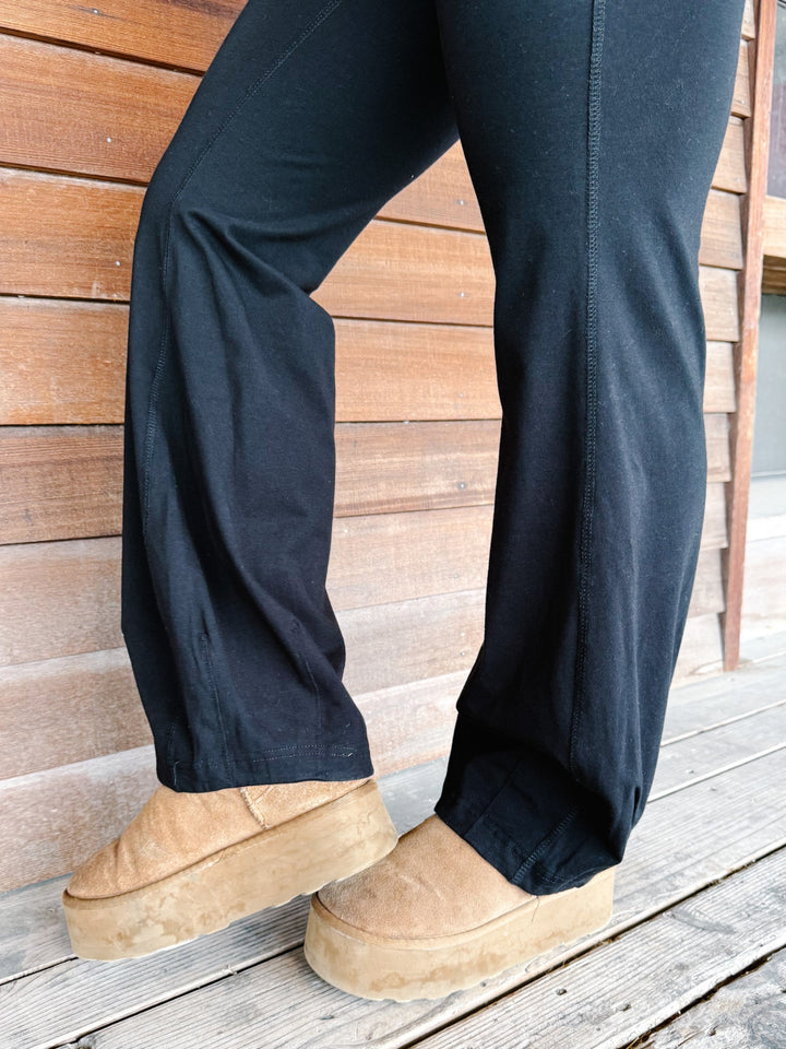 A person wearing black wide-leg harem pants with a drawstring waist, paired with a leopard-print sweatshirt and tan platform shoes, standing in front of a rustic wooden wall.