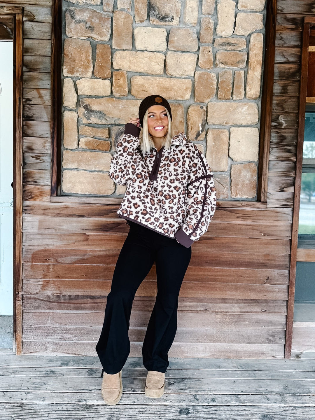 A person wearing black wide-leg harem pants with a drawstring waist, paired with a leopard-print sweatshirt and tan platform shoes, standing in front of a rustic wooden wall.