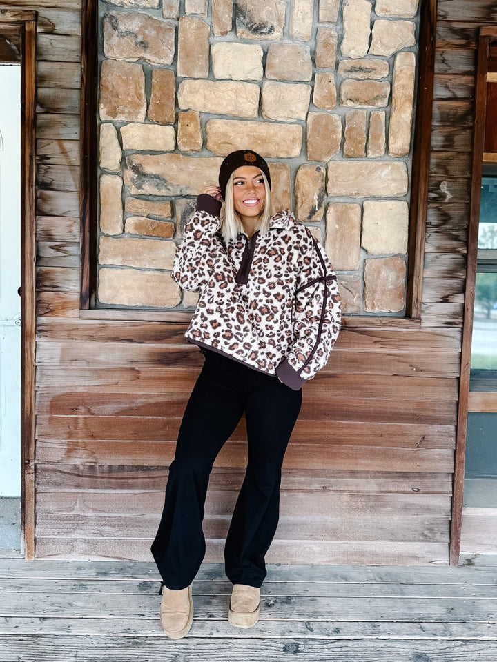 A person wearing black wide-leg harem pants with a drawstring waist, paired with a leopard-print sweatshirt and tan platform shoes, standing in front of a rustic wooden wall.