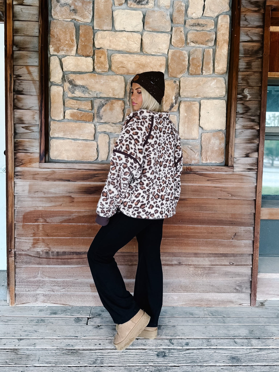 A person wearing black wide-leg harem pants with a drawstring waist, paired with a leopard-print sweatshirt and tan platform shoes, standing in front of a rustic wooden wall.