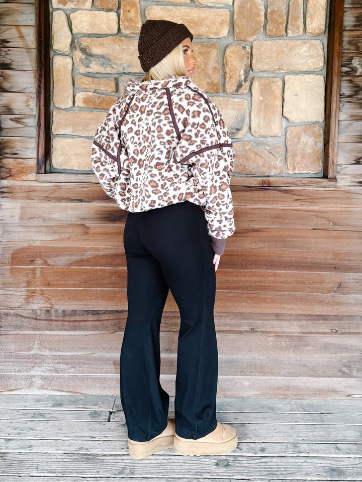A person wearing black wide-leg harem pants with a drawstring waist, paired with a leopard-print sweatshirt and tan platform shoes, standing in front of a rustic wooden wall.