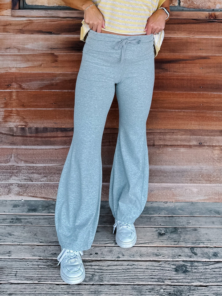 A person wearing light heather gray drawstring flare sweatpants, a yellow and heather oatmeal striped top, and white sneakers, standing against a rustic wooden wall and deck.