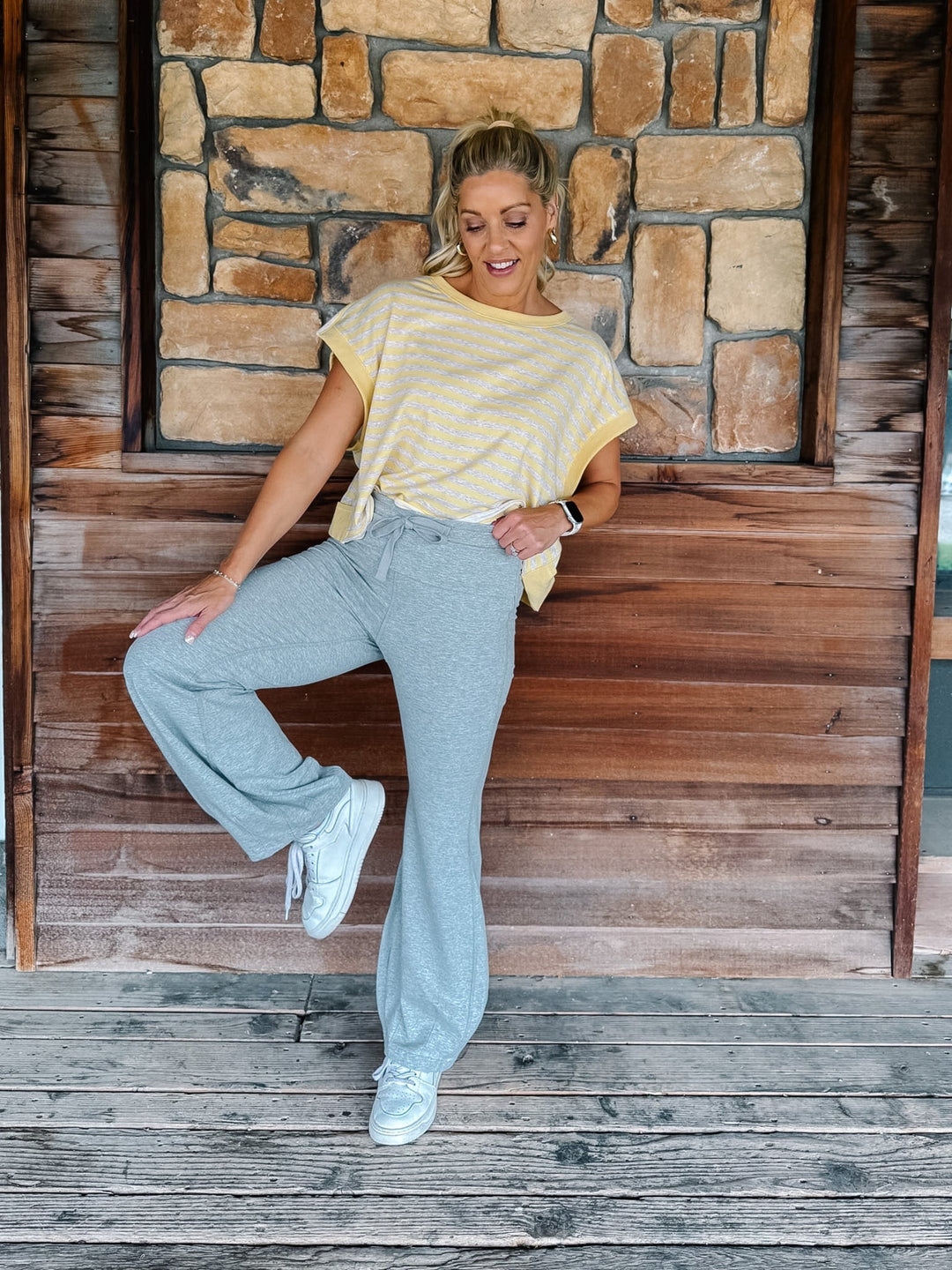 A person wearing light heather gray drawstring flare sweatpants, a yellow and heather oatmeal striped top, and white sneakers, standing against a rustic wooden wall and deck.