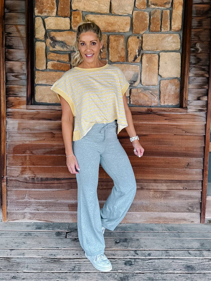 A person wearing light heather gray drawstring flare sweatpants, a yellow and heather oatmeal striped top, and white sneakers, standing against a rustic wooden wall and deck.