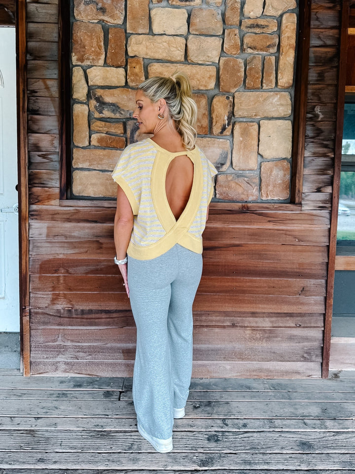 A person wearing light heather gray drawstring flare sweatpants, a yellow and heather oatmeal striped top, and white sneakers, standing against a rustic wooden wall and deck.