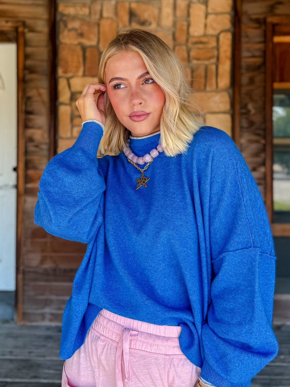 A blonde woman wearing a vibrant blue oversized turtleneck sweater and light pink sweatpants, standing in front of a rustic stone and wood building.