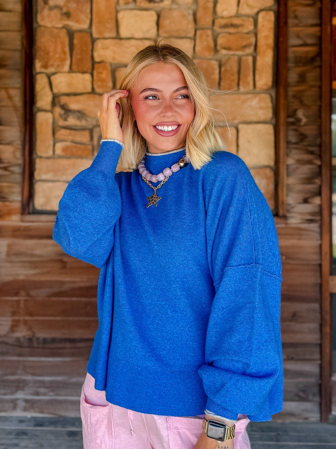 A smiling blonde woman wearing an oversized bright blue knit sweater and light pink pants, standing in front of a rustic stone and wood wall.