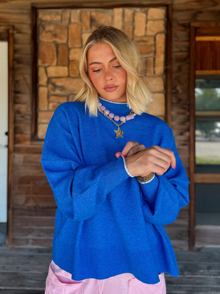 A blonde woman wearing a bright royal blue knit sweater and pink pants, standing on a wooden porch in front of a stone wall.