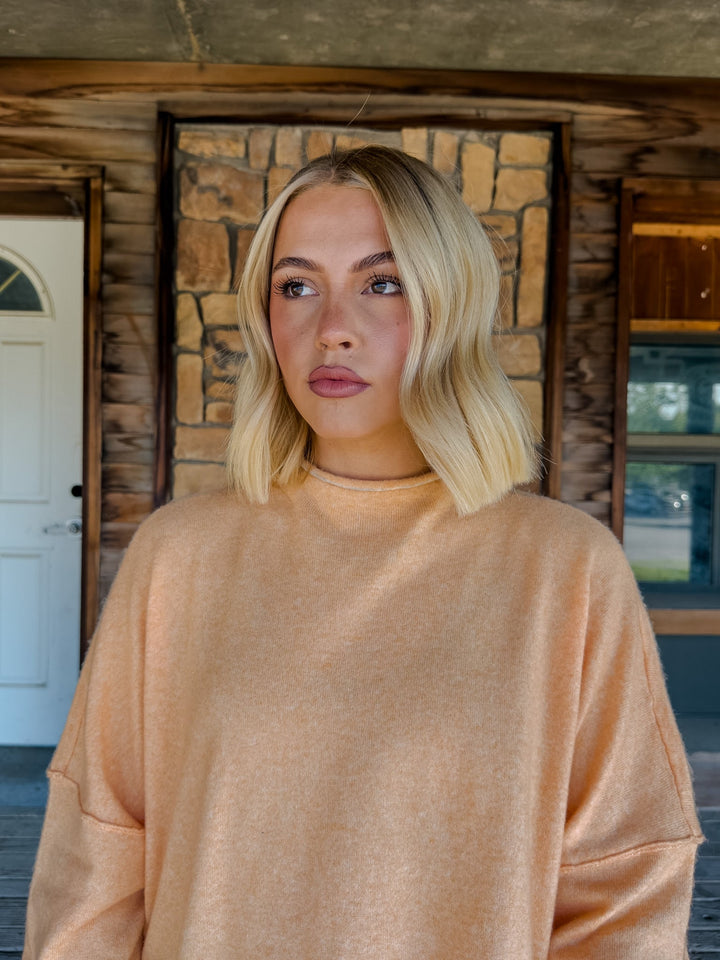 A blonde woman wearing a soft peach-colored knit sweater, standing in front of a rustic log cabin wall with a stone accent panel.