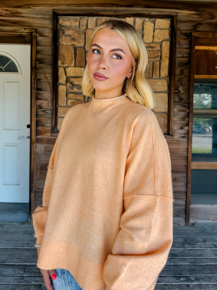 A blonde woman wearing a loose, oversized peach-colored knit sweater, standing on a wooden porch in front of a rustic stone and wood wall.