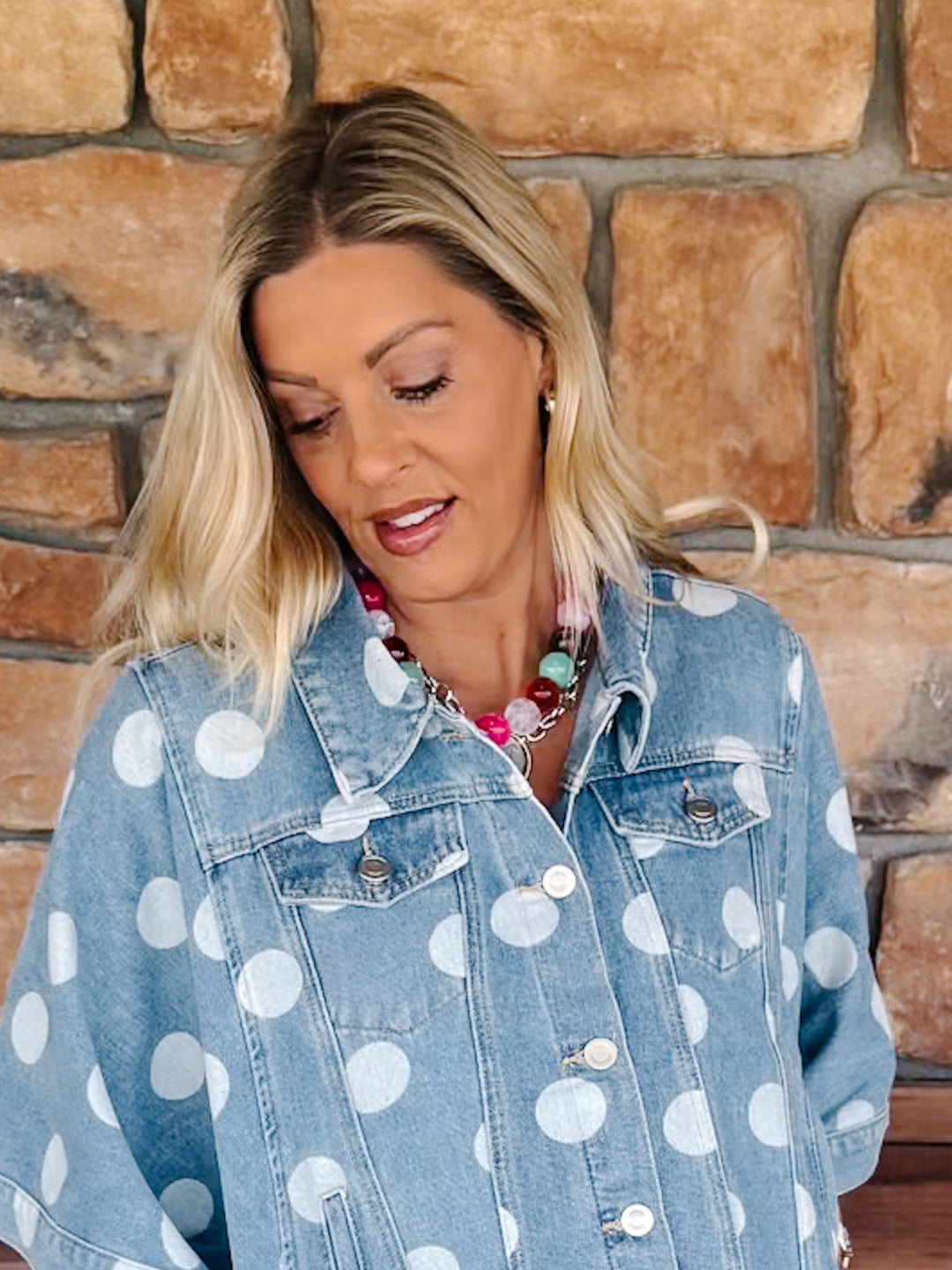 A woman with blonde hair wearing a light blue denim jacket with large white polka dots, standing in front of a rustic stone wall.