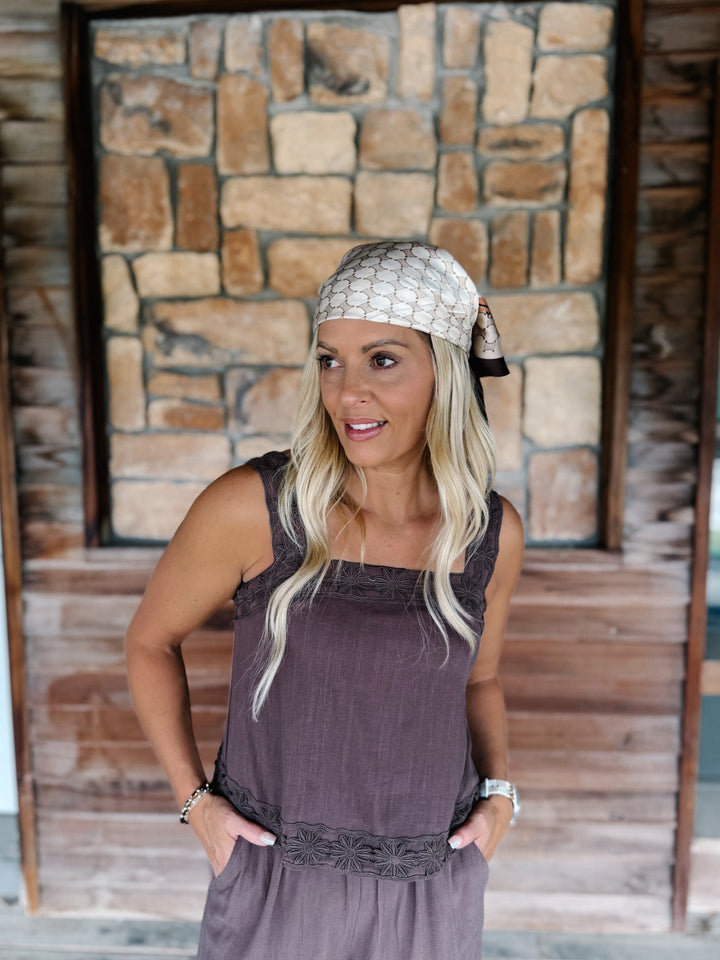 A woman with blonde hair wearing a mauve sleeveless top, a matching headscarf, and a matching skirt, standing in front of a rustic stone and wood wall.
