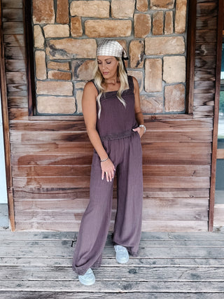 A woman wearing a sleeveless mocha wide-leg jumpsuit, a beige patterned headwrap, and white sneakers, standing on a wooden deck in front of a rustic stone and wood wall.