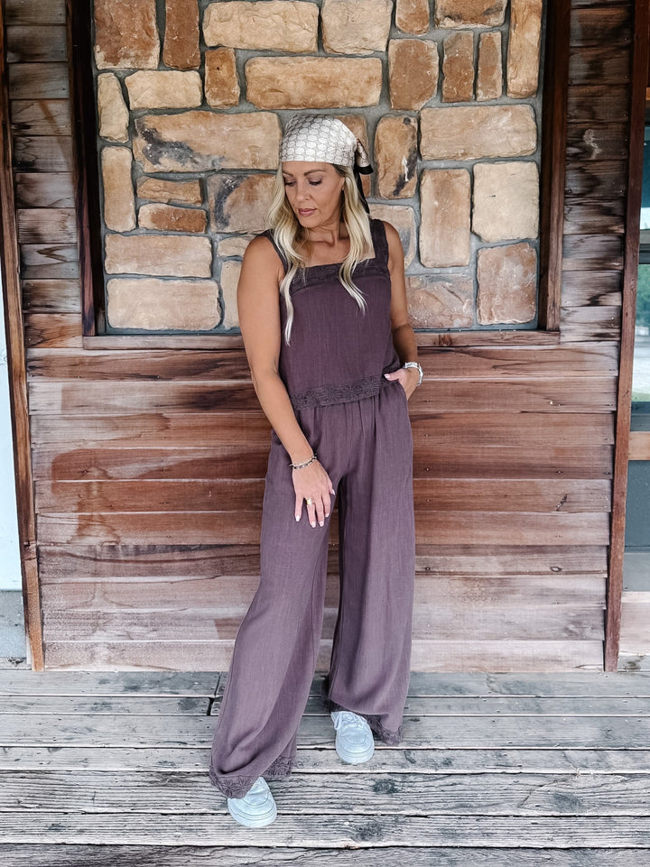 A woman wearing a sleeveless mocha wide-leg jumpsuit, a beige patterned headwrap, and white sneakers, standing on a wooden deck in front of a rustic stone and wood wall.