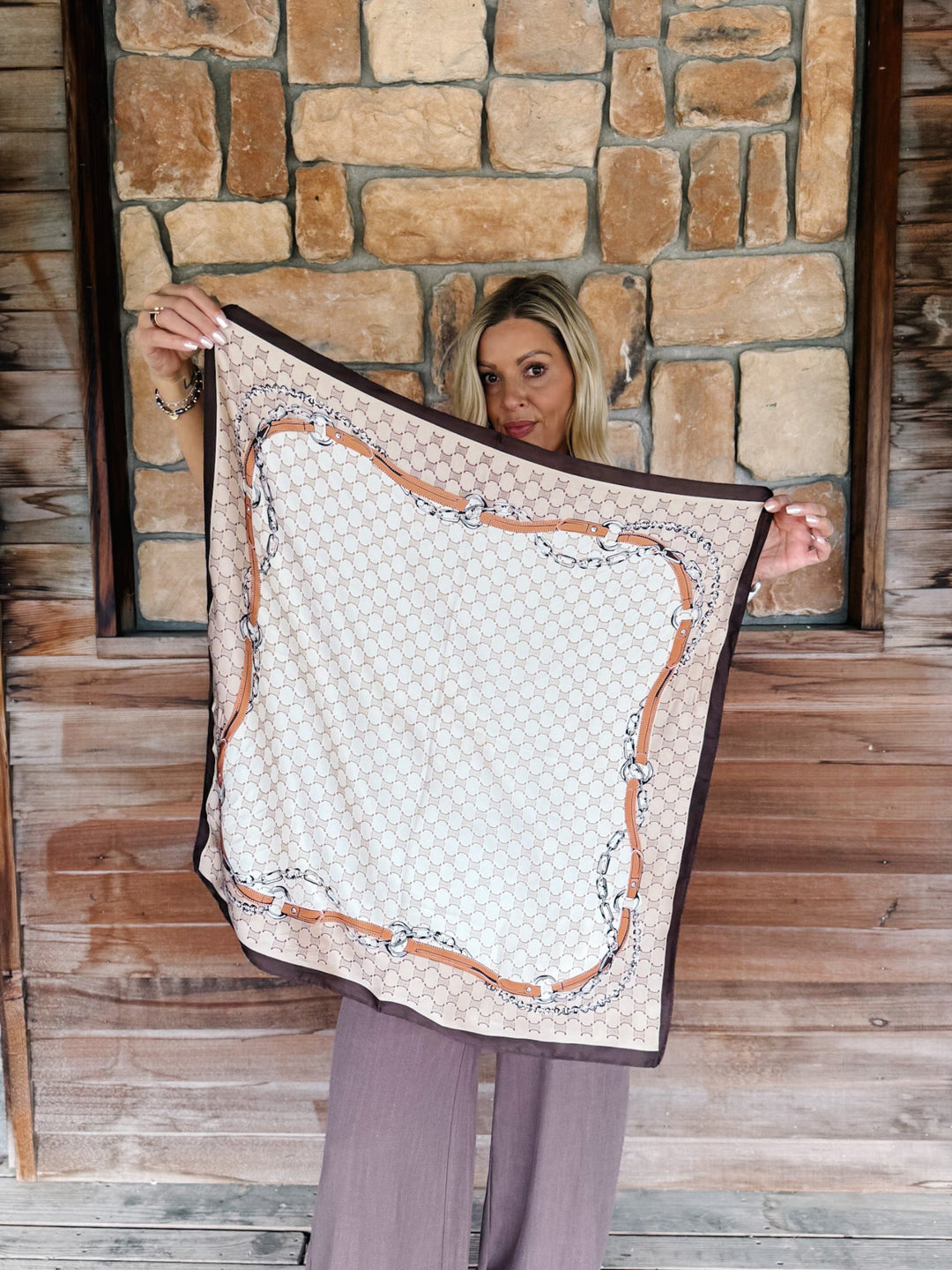 A woman holds up a square scarf featuring a beige and white geometric pattern with a brown and silver chain-link border, set against a rustic stone and wood wall background.