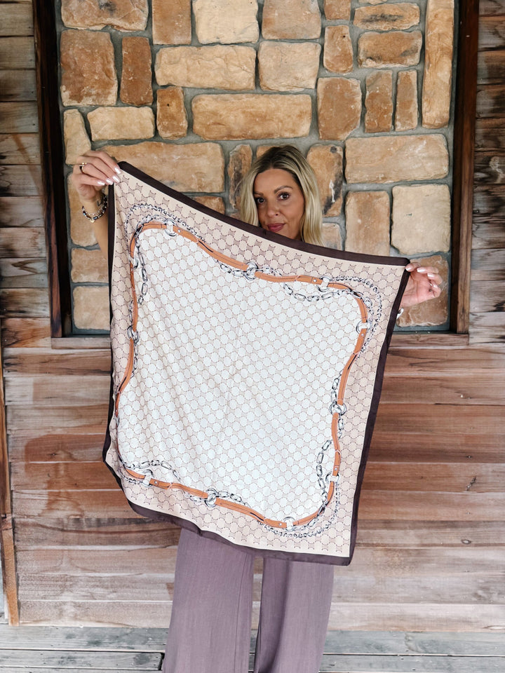 A woman holds up a square scarf featuring a beige and white geometric pattern with a brown and silver chain-link border, set against a rustic stone and wood wall background.