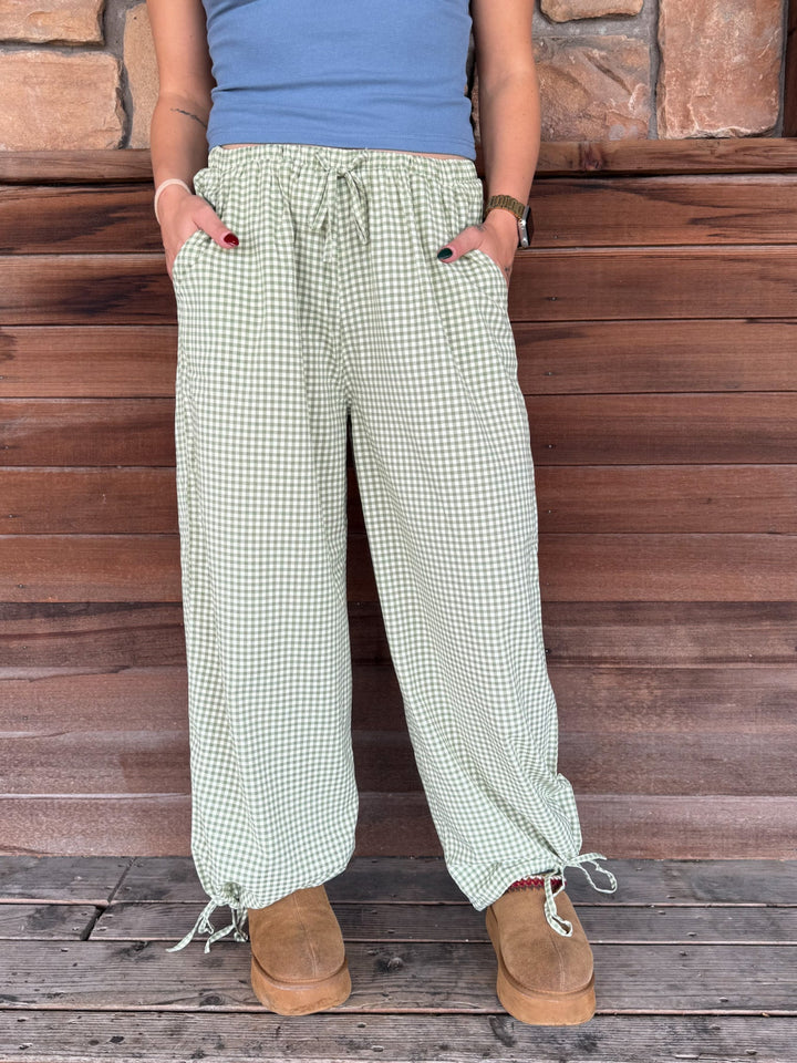 A person wearing loose, wide-leg gingham pants in a light green and white check pattern, paired with a blue top and brown boots, standing against a rustic wooden wall.