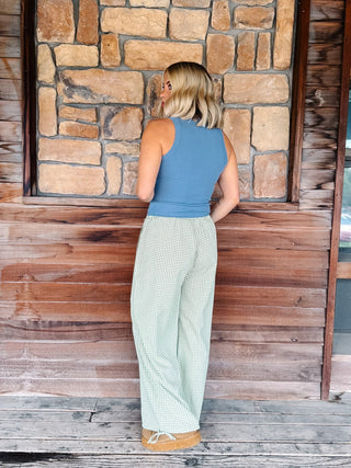 A woman stands with her back to the camera wearing a slate blue sleeveless top and light green and white gingham wide-leg pants, posing in front of a rustic stone and wood wall.