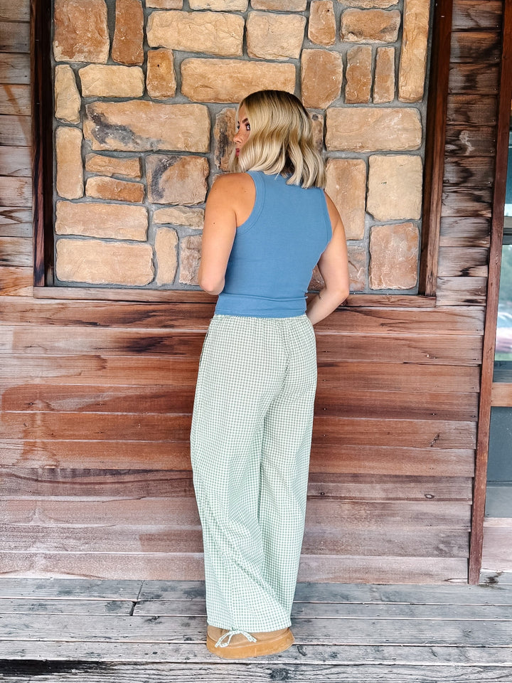 A woman stands with her back to the camera wearing a slate blue sleeveless top and light green and white gingham wide-leg pants, posing in front of a rustic stone and wood wall.