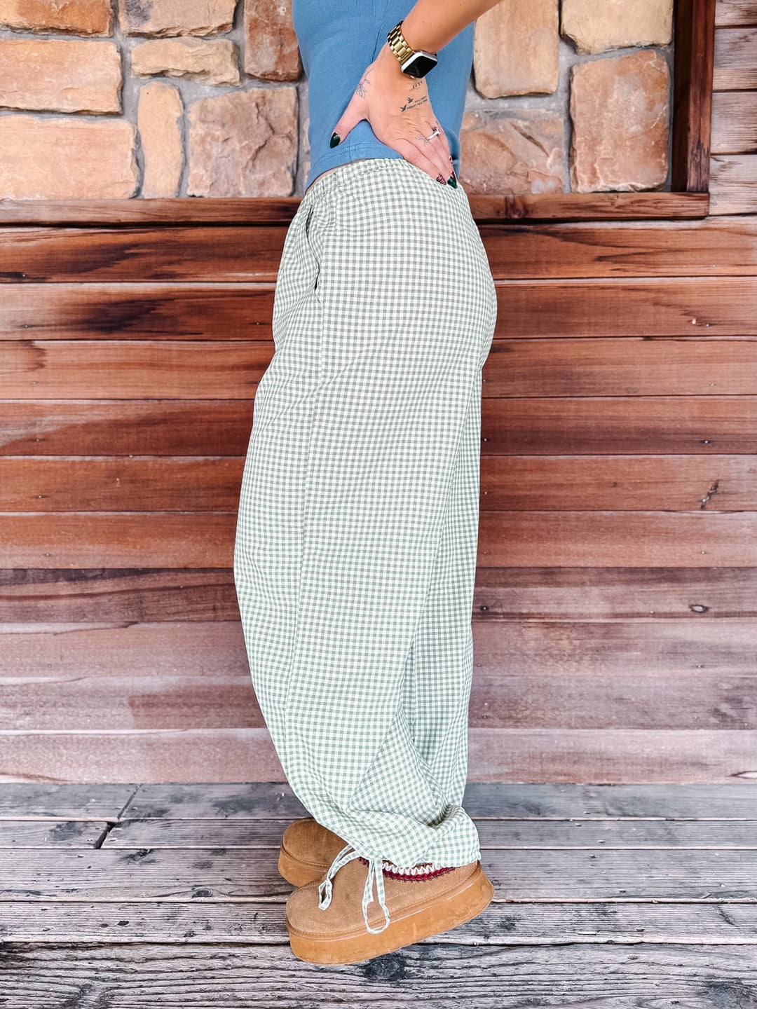 A person shown from the waist down wearing loose, wide-leg checkered pants in a light green and white gingham pattern, paired with a blue top and tan boots, standing against a rustic wooden and stone wall.