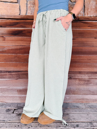 A person wearing light green and white gingham wide-leg drawstring pants, paired with a blue top and tan boots, standing in front of a rustic wooden wall.