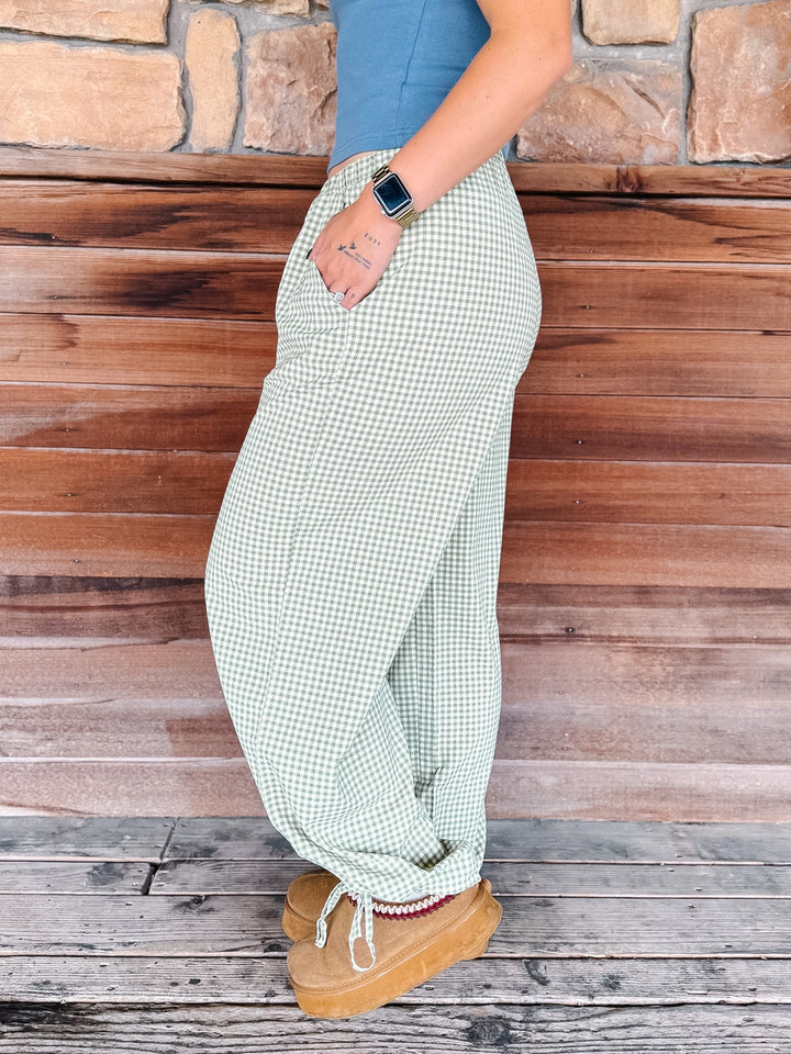 A person wearing light green and white gingham wide-leg pants, a blue top, and tan boots, standing against a rustic wooden plank wall.
