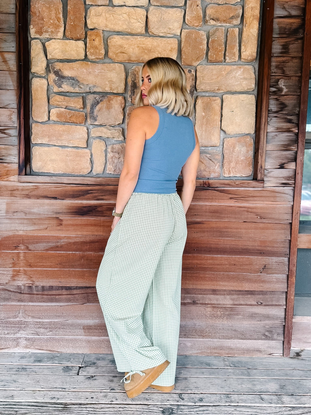 A woman stands on a wooden porch in front of a stone wall, wearing a sleeveless slate blue top and light sage green wide-leg pants with a small check pattern.