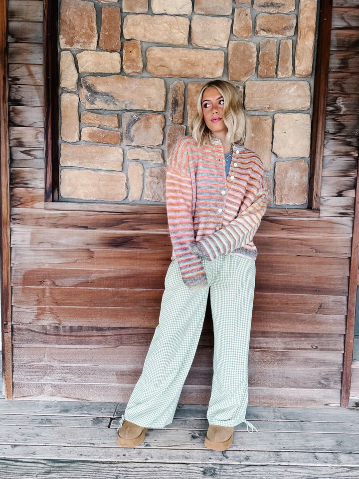 A woman stands on a rustic wooden porch in front of a stone wall, wearing a striped button-front cardigan in peach, orange, and gray tones, paired with light sage green wide-leg pants and tan boots.
