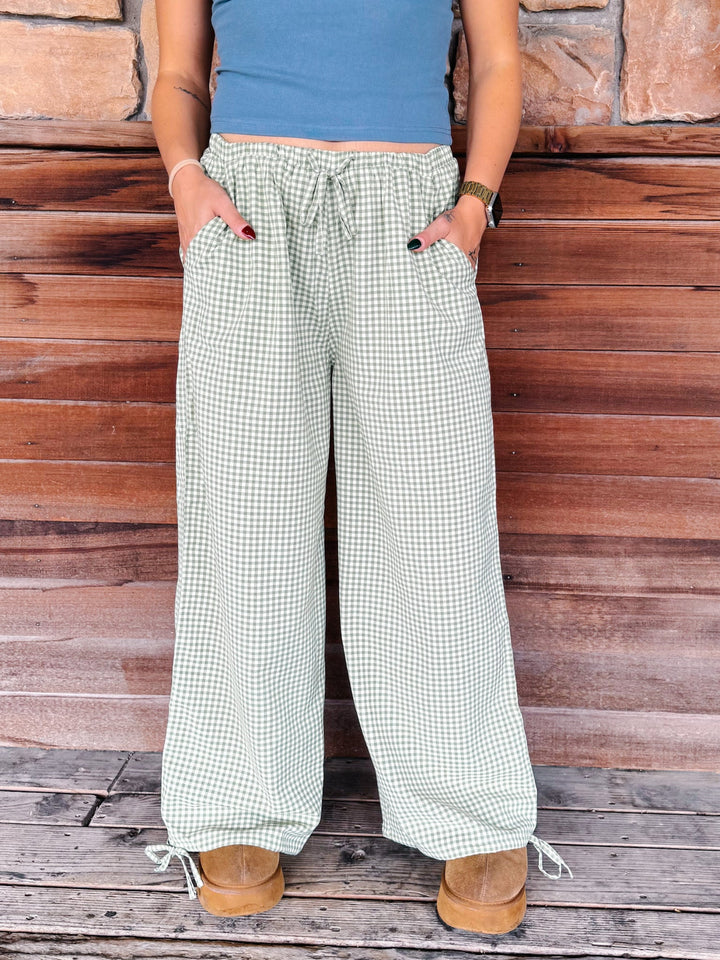 A person wearing wide-leg drawstring pants with a green and white gingham pattern, standing against a rustic wooden plank wall.