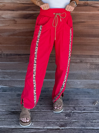 Red wide-leg pants with beige and black patterned side stripes, modeled against a rustic wooden plank background.