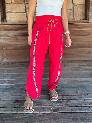 A person wearing bright red wide-leg pants with beige patterned side stripes and a beige drawstring waist, standing on a wooden deck in front of a stone wall.