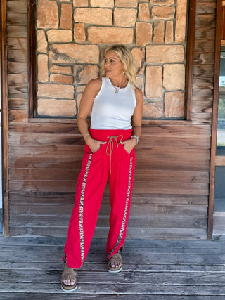 A woman stands on a wooden porch wearing bright red drawstring pants with white lettering down the sides, a white sleeveless top, and brown slide sandals, in front of a stone and wood wall.
