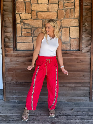 A woman stands on a wooden porch in front of a stone wall, wearing bright red drawstring pants with a patterned side stripe, a white sleeveless top, and brown sandals.