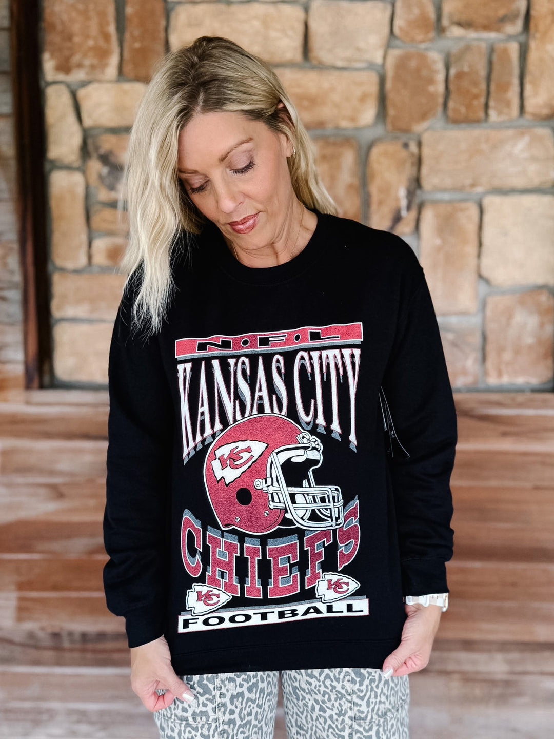 A woman wearing a black long-sleeve crewneck sweatshirt with a red and white Kansas City Chiefs graphic, standing in front of a stone wall.