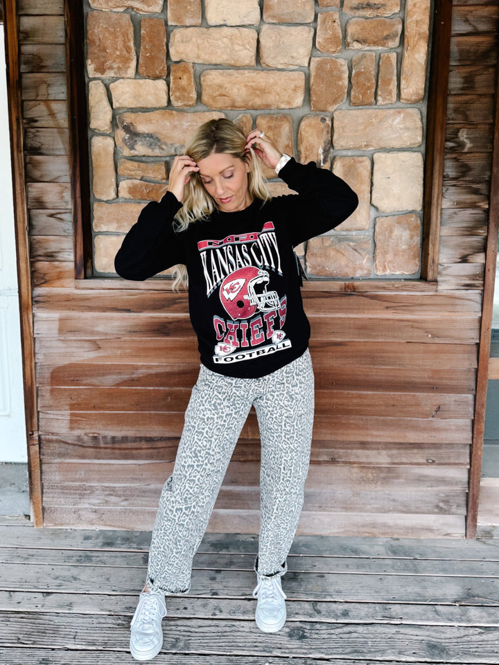 A woman stands on a wooden deck in front of a stone wall, wearing a black crewneck sweatshirt with a red and white &