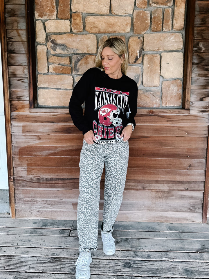 A woman stands on a wooden deck in front of a stone wall, wearing a black long-sleeve graphic sweatshirt with a red and white &