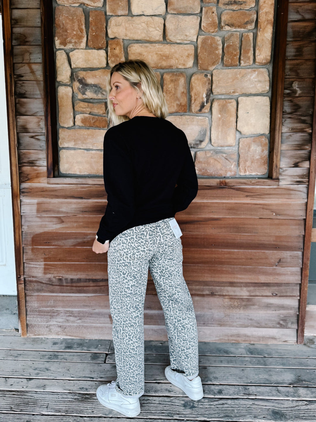 A woman stands on a wooden porch wearing a black long-sleeve top, gray and white patterned pants, and white sneakers, photographed from the back.