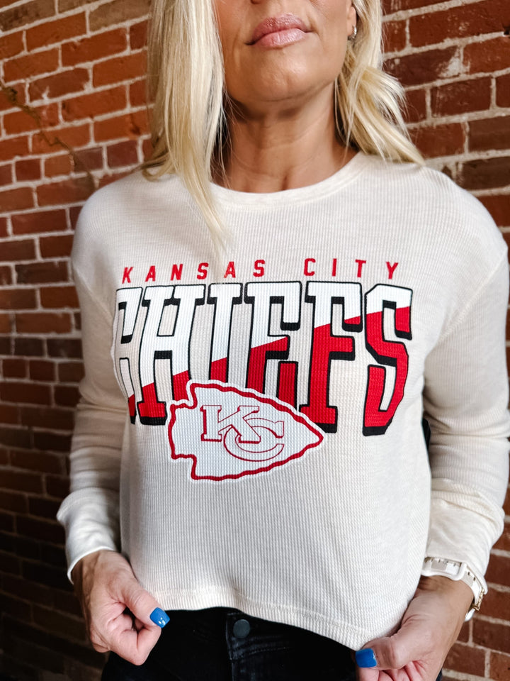 A woman wearing a cream ribbed long-sleeve crewneck sweatshirt with a large Kansas City Chiefs graphic on the front, standing against a red brick wall.